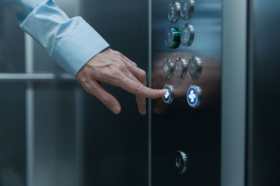 A close-up of a person's hand pressing the button to operate an elevator lift inside a residential building. The individual wears a light blue long-sleeved shirt, with visible veins on the hand. The e
