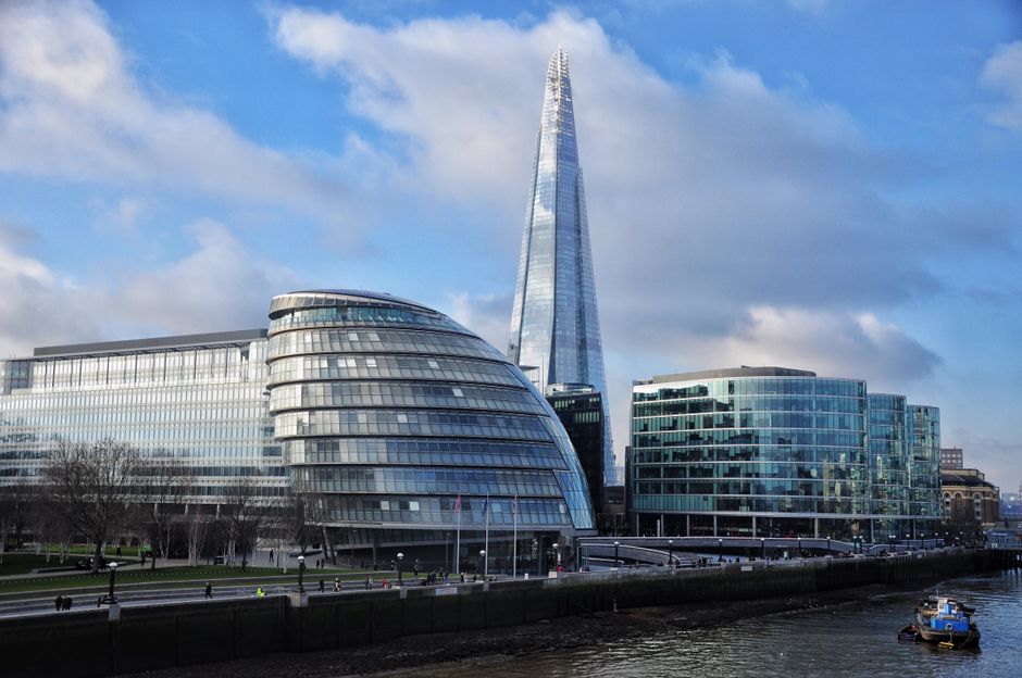 A view of the Shard skyscraper in London, standing tall against a cloudy sky, with surrounding buildings including a mix of modern and older architectural styles. In the foreground, a river runs benea