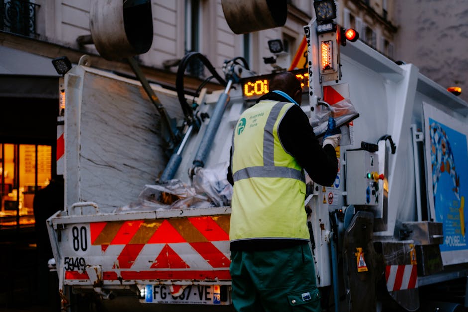 Two men engaged in a loading process of waste disposal for house removals in Southwark. One man, dressed in a black jacket and wearing yellow gloves, is placing a cardboard box filled with waste into 